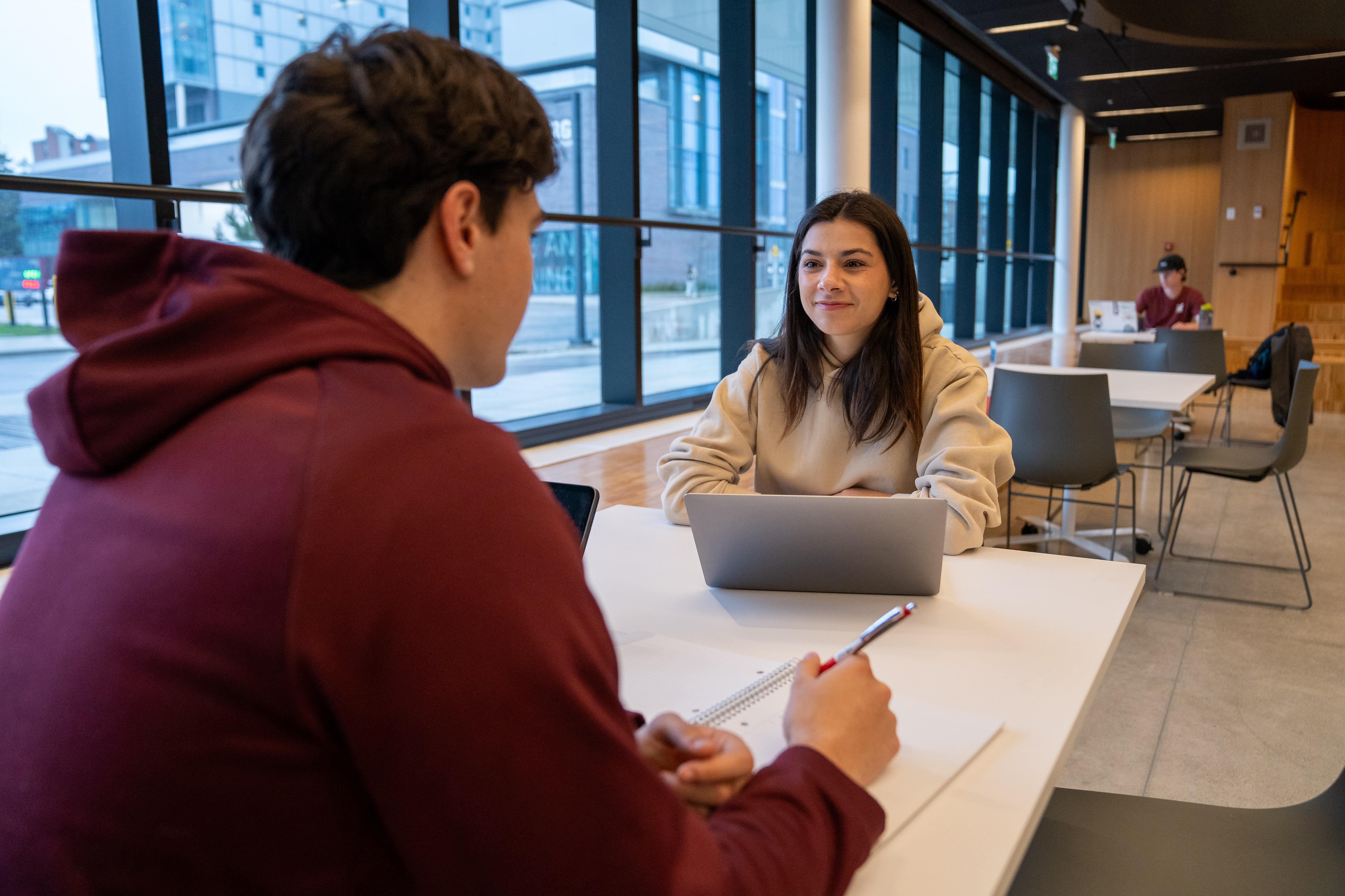 Two students sitting at a table in conversation with a laptop in between.